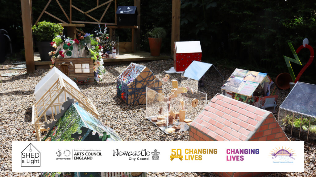 This is a colour photograph of a garden scene with a gravel floor and hedge surrounding it. In the garden are twelve miniature sheds with a range of artistic designs. In the back of the frame, two of the sheds are sat on a covered wooden decking. Overlaying the image at the bottom is a banner containing six logos.