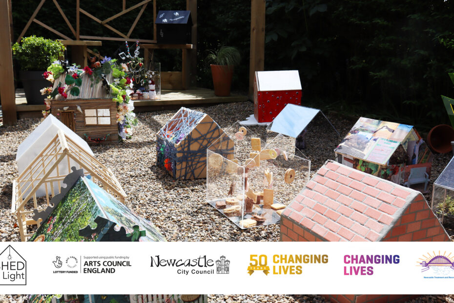 This is a colour photograph of a garden scene with a gravel floor and hedge surrounding it. In the garden are twelve miniature sheds with a range of artistic designs. In the back of the frame, two of the sheds are sat on a covered wooden decking. Overlaying the image at the bottom is a banner containing six logos.