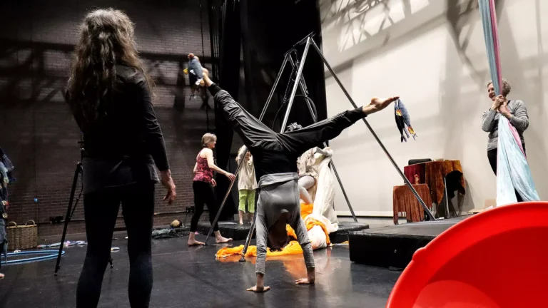 Performers are using a variety of aerial kit in a theatre. The person in the centre is doing handstand splits.