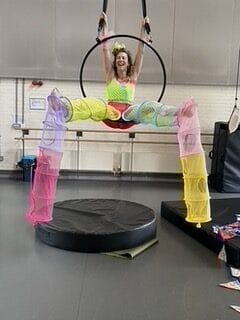 A female aerialist performing on an aerial hoop. The aerialist is laughing and wearing a colourful costume made of recycled storage nets