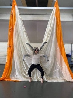 A woman with sunglasses posing in front of two large pieces of cream and orange fabrics, spreading like butterfly wings